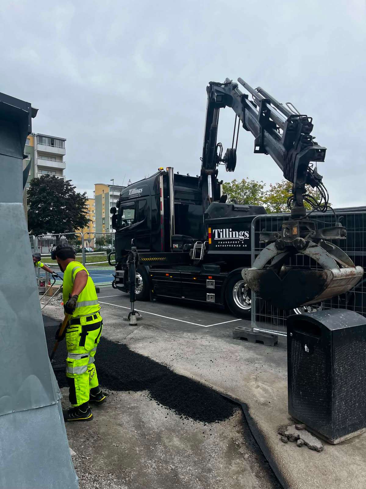 Kran med gripskopa lossar asfalt, förare i varselkläder